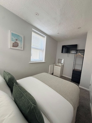 Quarto com cama, cômoda, espelho, televisão e janela com persianas.