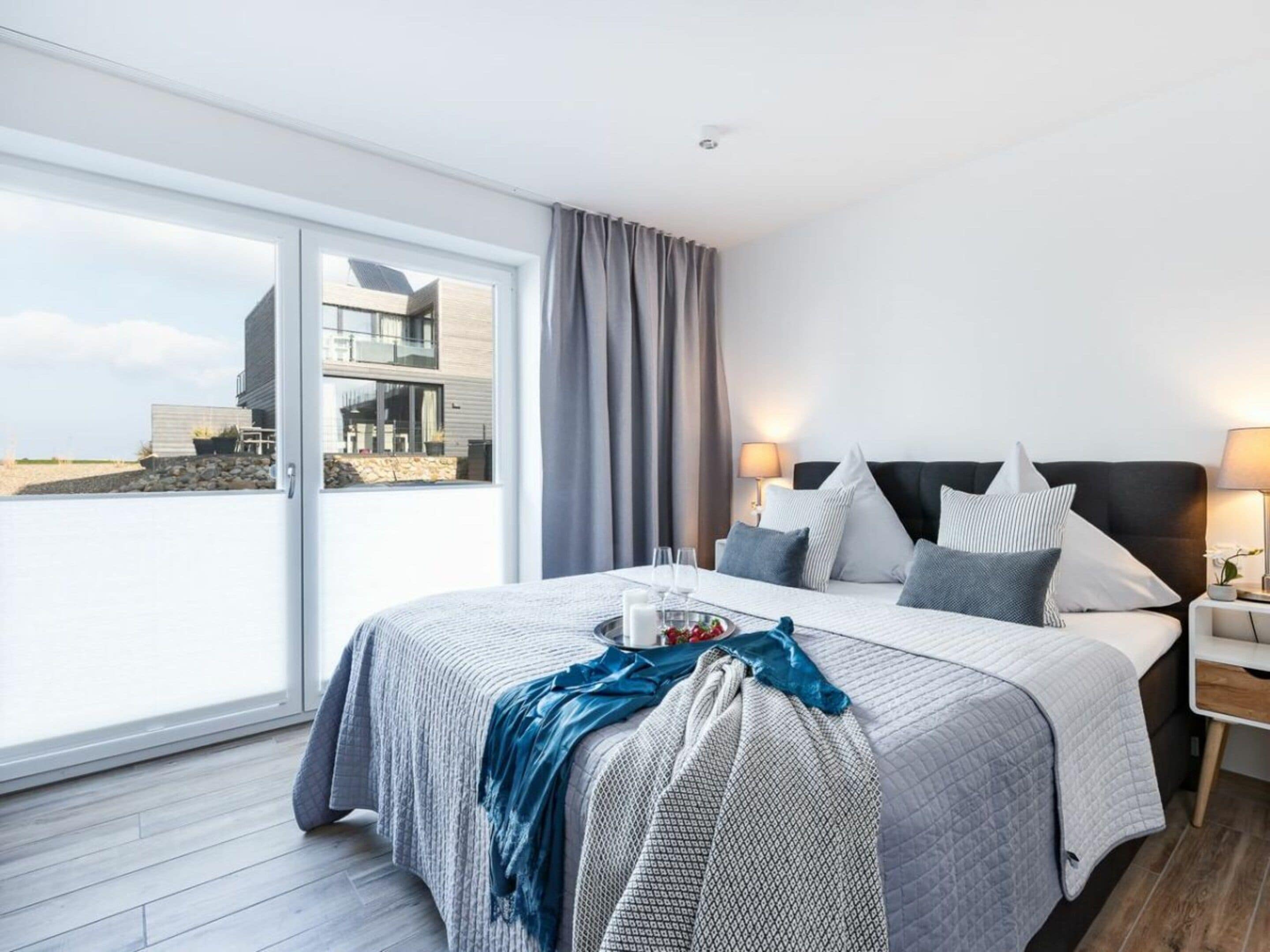 A modern hotel room with a large bed, a nightstand, a lamp, and a view of the beach.