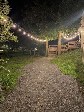 Un sendero de grava conduce a una estructura de madera con barandilla, rodeada de follaje y luces colgantes.