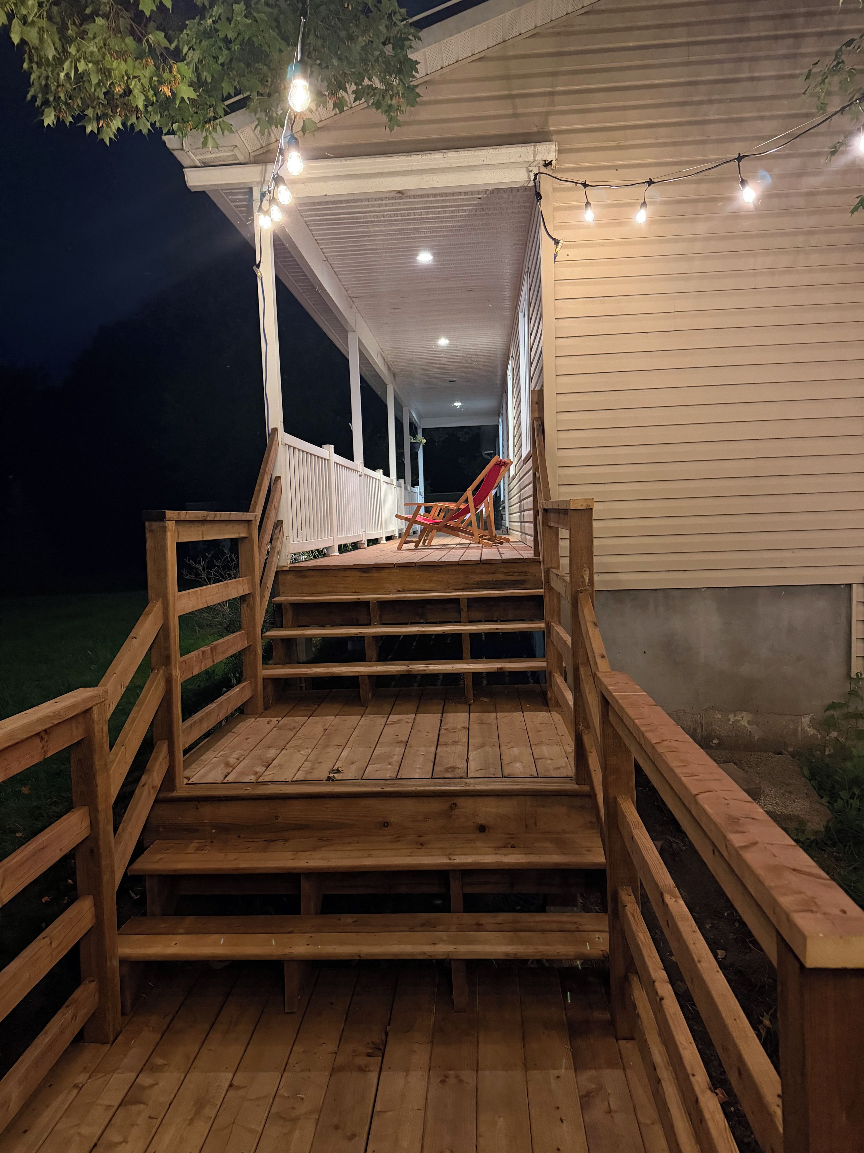 A wooden deck with steps leading to a covered porch, a red chair, and string lights.