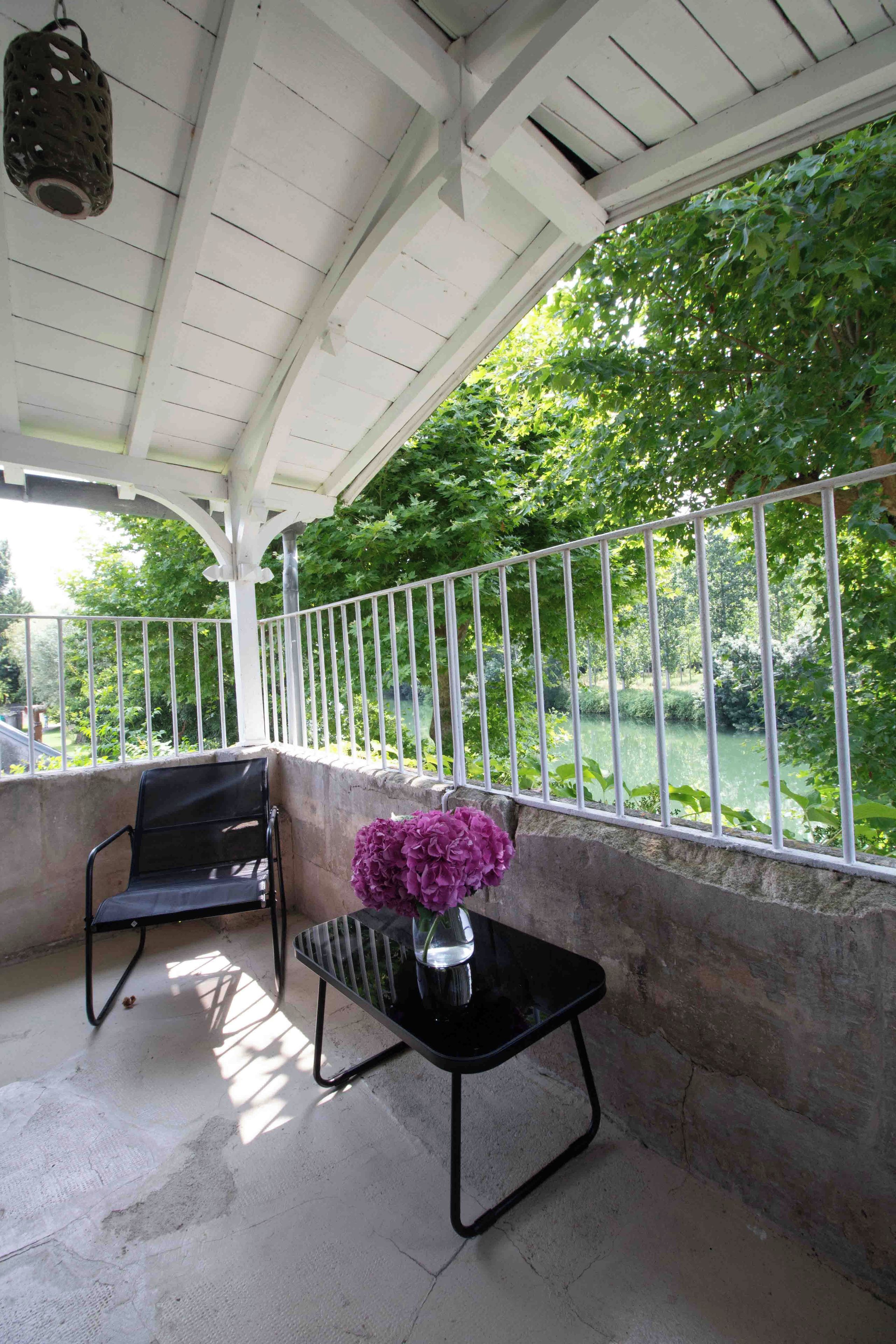 A covered outdoor area with a table and chairs, a metal railing, and a view of a green landscape.