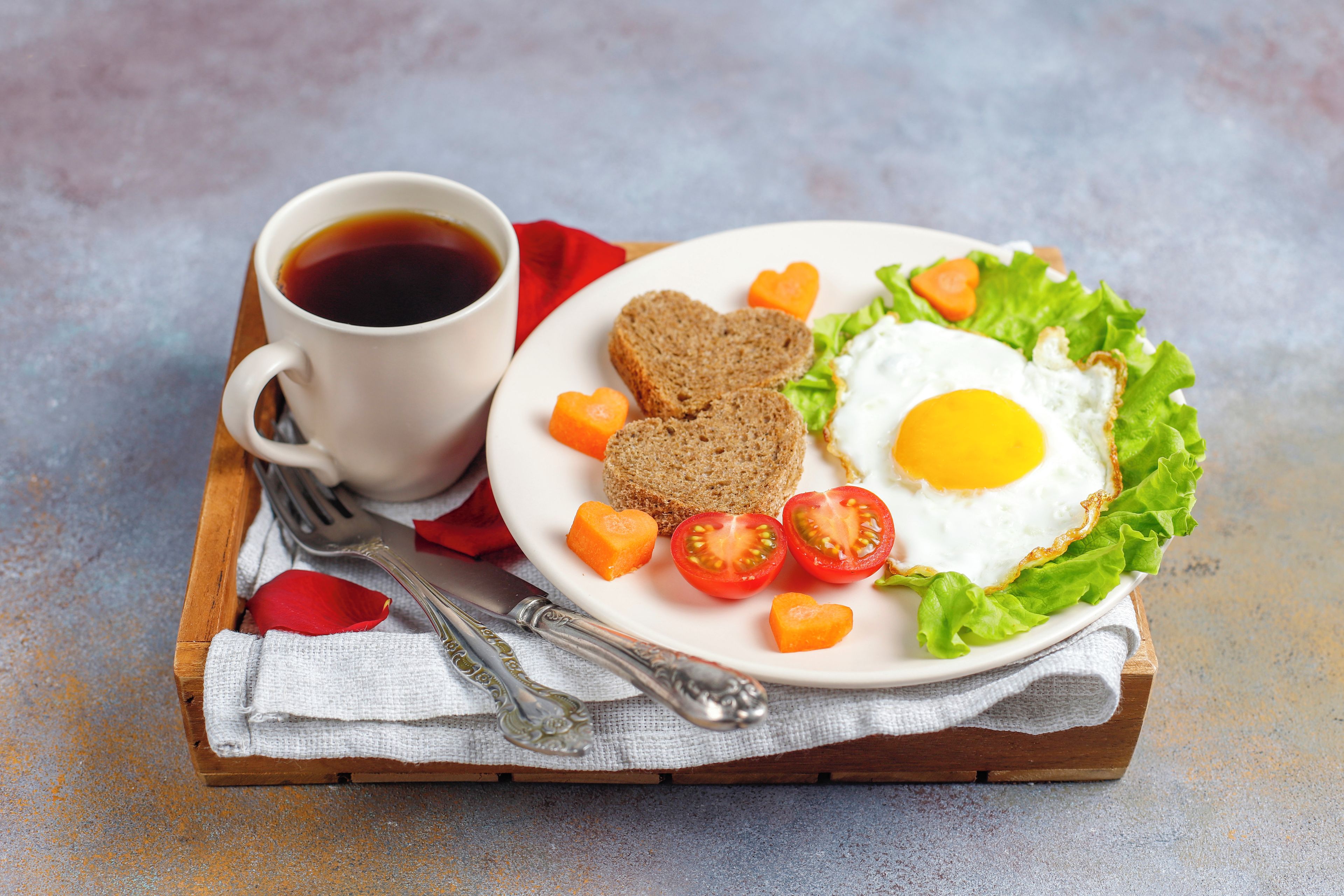 A plate with a fried egg, tomato slices, carrot heart shapes, and bread on a white plate, accompanied by a cup of coffee on a wooden tray.