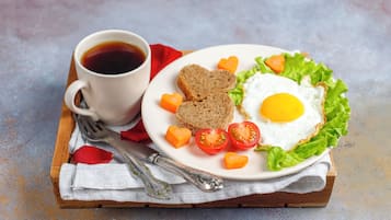 A plate with a fried egg, tomato slices, carrot heart shapes, and bread on a white plate, accompanied by a cup of coffee on a wooden tray.