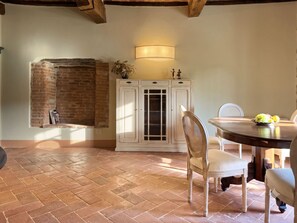A dining area with a brick fireplace, white cabinetry, and a round table with chairs. - Casale Tre Dame - Appartamento L Aretino (Castiglion Fiorentino)