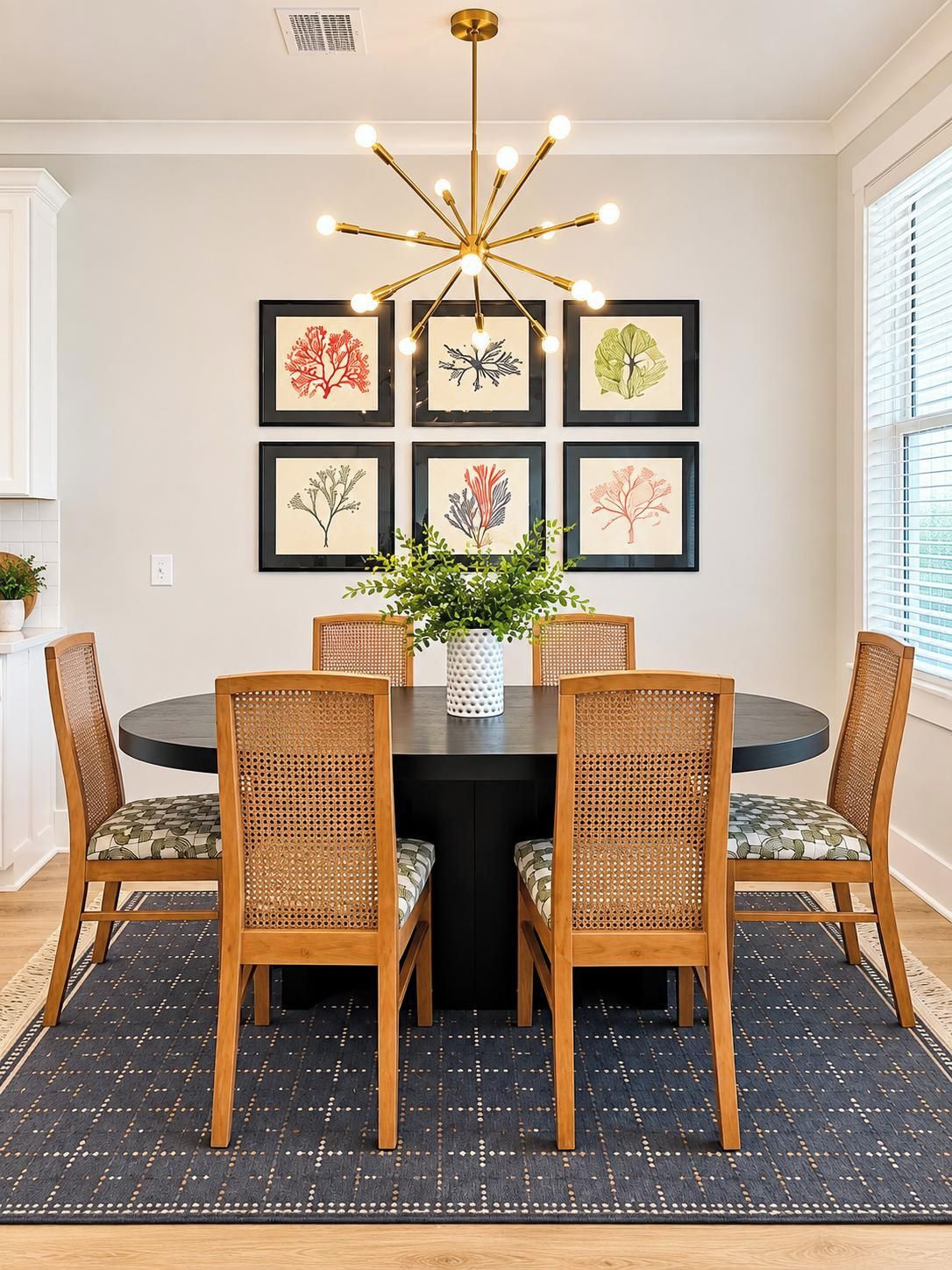 A dining area with a round table, four chairs, a central vase with greenery, and framed botanical prints on the wall.
