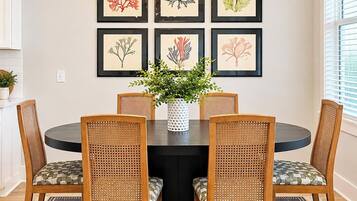 A dining area with a round table, four chairs, a central vase with greenery, and framed botanical prints on the wall.