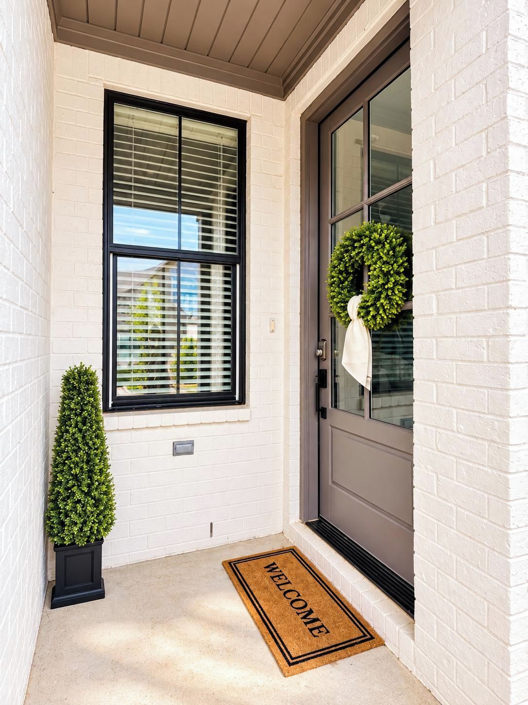 A front door with a 'WELCOME' mat, a potted plant, a wreath, and a window with blinds.