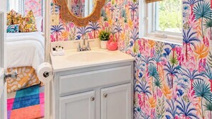 Une salle de bain avec un papier peint à motifs floraux, un miroir avec un cadre doré, un lavabo avec un plan de travail blanc et une fenêtre avec un store en bambou.