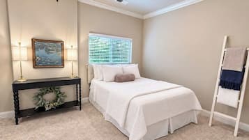 Un dormitorio con cama, una mesita de consola con un arreglo floral, un cuadro y una ventana con persianas.