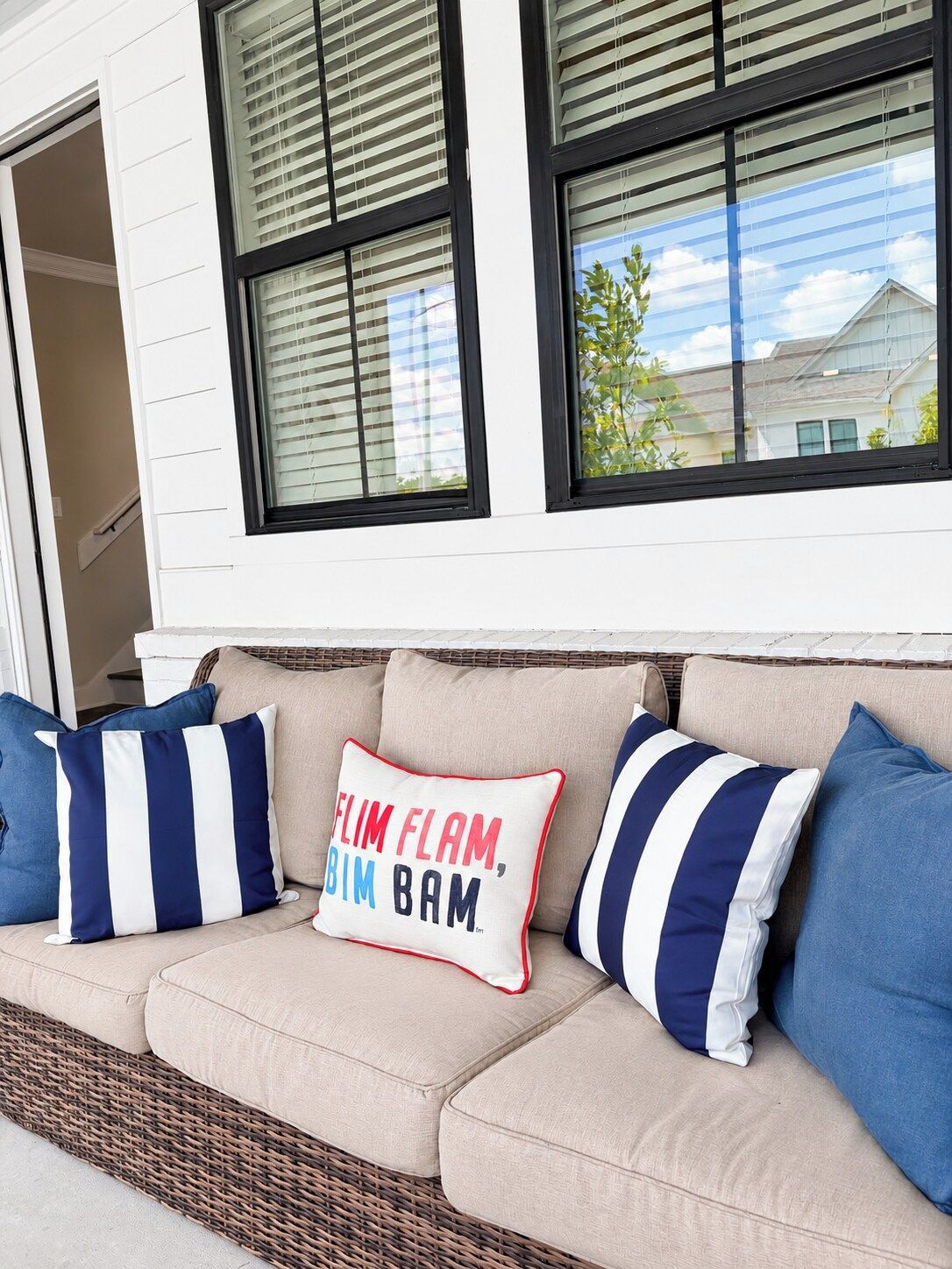 A beige outdoor sofa with striped pillows, positioned in front of a white building with black-framed windows.