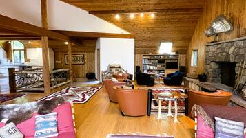 A cozy cabin interior with a stone fireplace, wooden beams, a dining area, and a living room furnished with sofas and a coffee table.