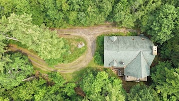 An aerial view of a house surrounded by dense forest, with a visible driveway leading to the entrance.