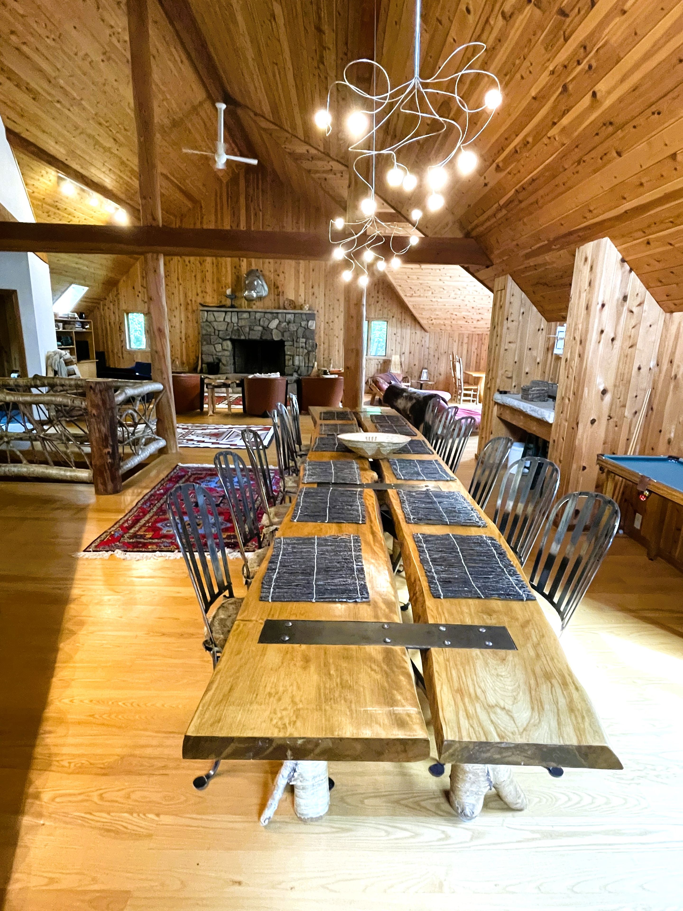 A wooden interior with a long dining table, chairs, a fireplace, and a hanging light fixture.