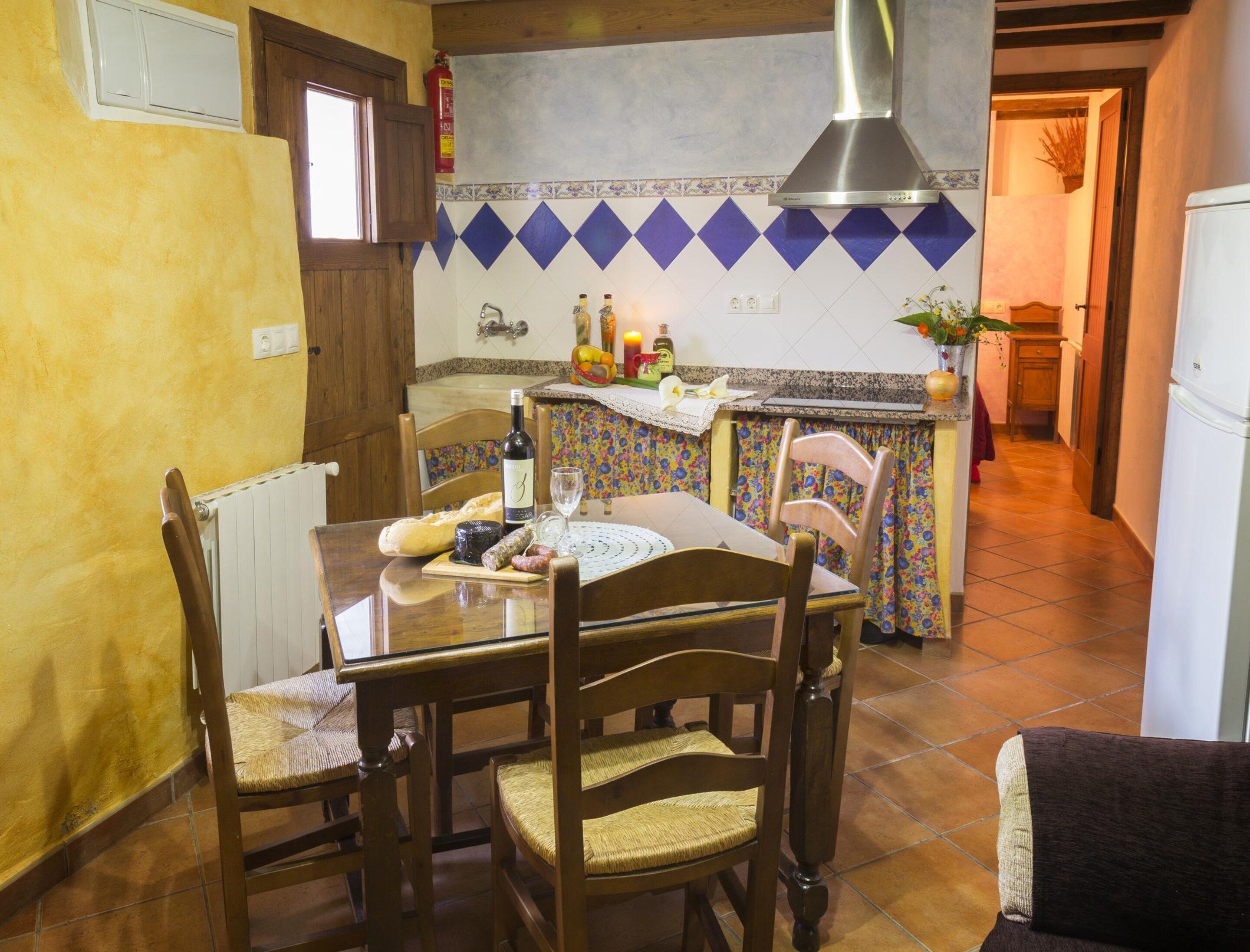 Comedor con una mesa lista para cuatro, sillas, un radiador, una cocina al fondo con un salpicadero de azulejos y un extintor de incendios montado en la pared.