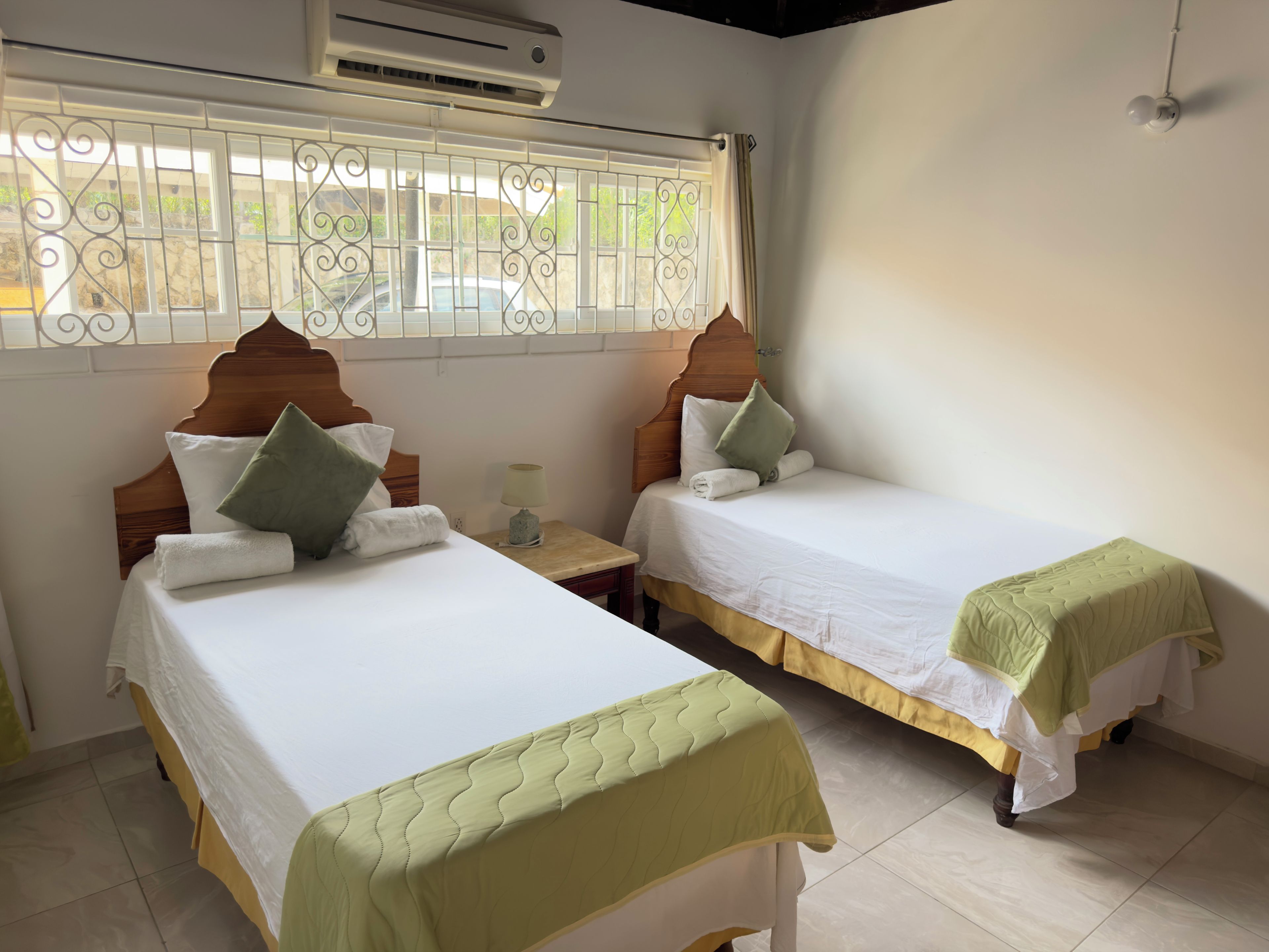 Quarto de hotel com duas camas, janela com grades decorativas, ar condicionado e mesa de cabeceira com abajur.