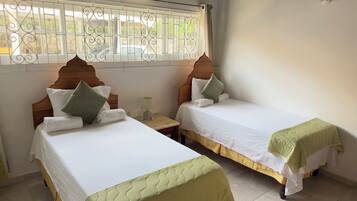 Quarto de hotel com duas camas, janela com grades decorativas, ar condicionado e mesa de cabeceira com abajur.