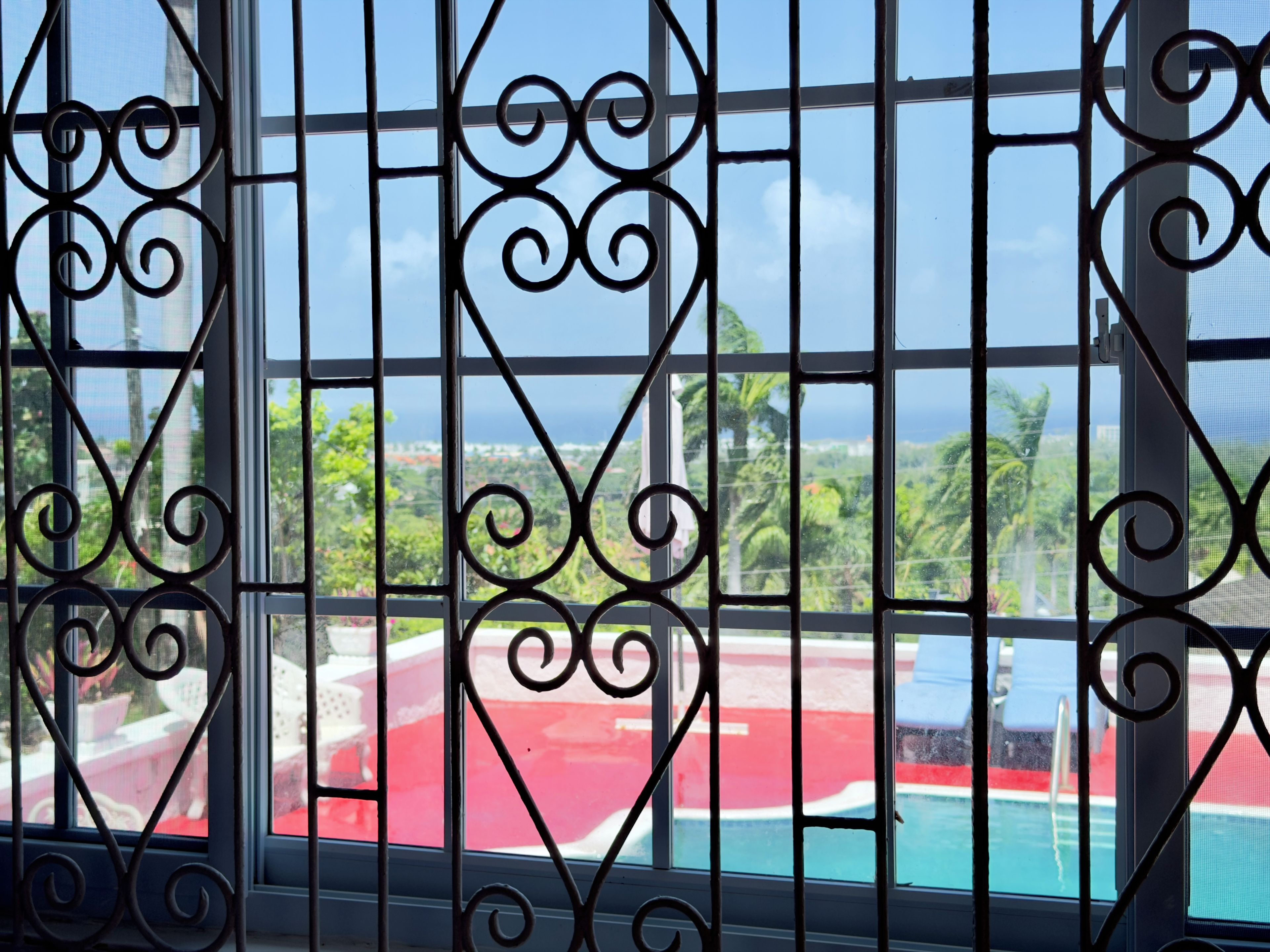 A window with decorative iron bars, a view of a swimming pool, and greenery outside.