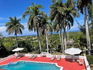A pool area with lounge chairs and umbrellas, surrounded by palm trees and a view of the landscape.