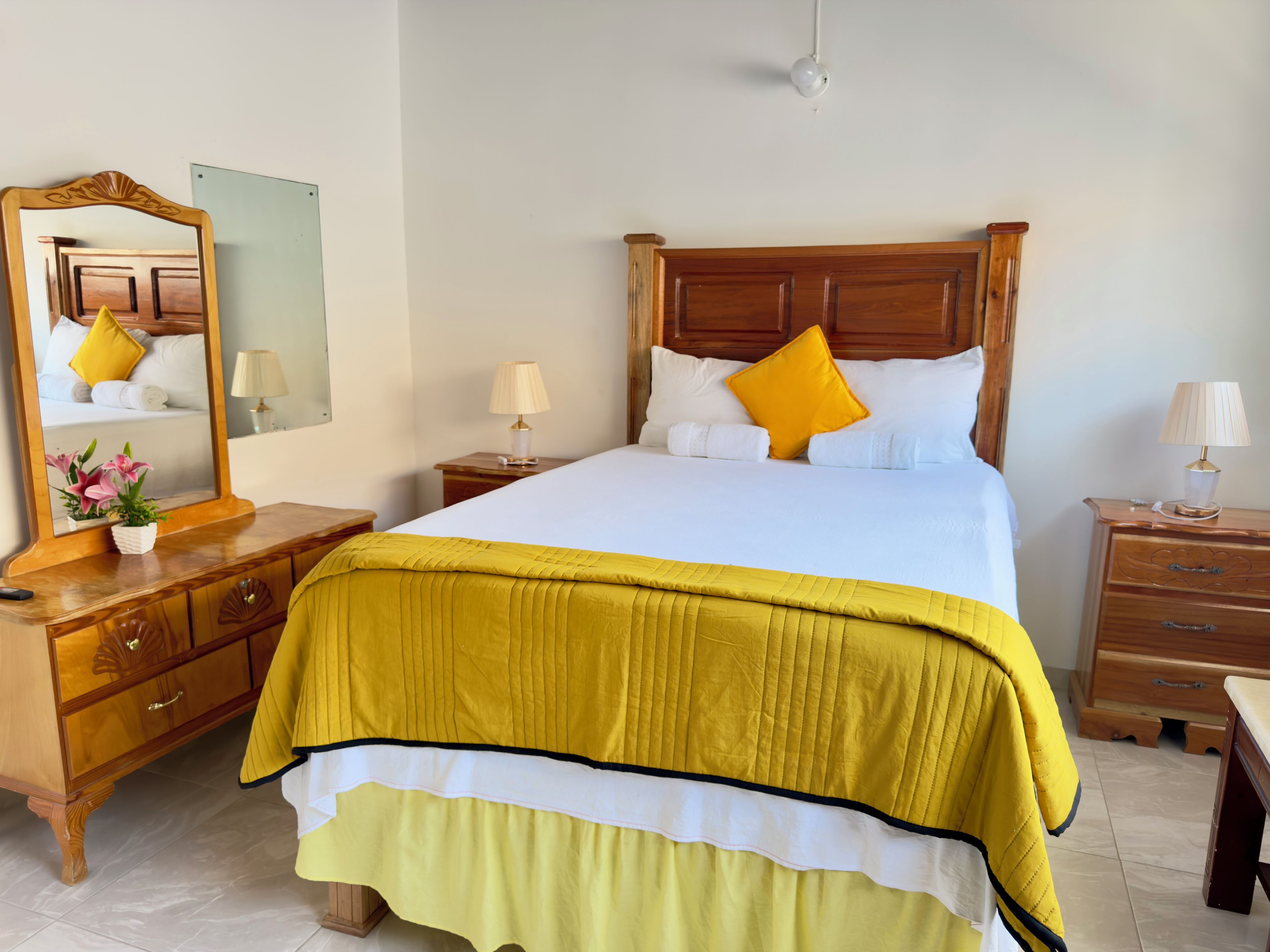 Una cama bien hecha con cabecera de madera, una mesita de noche con lámpara y un espejo en la pared.