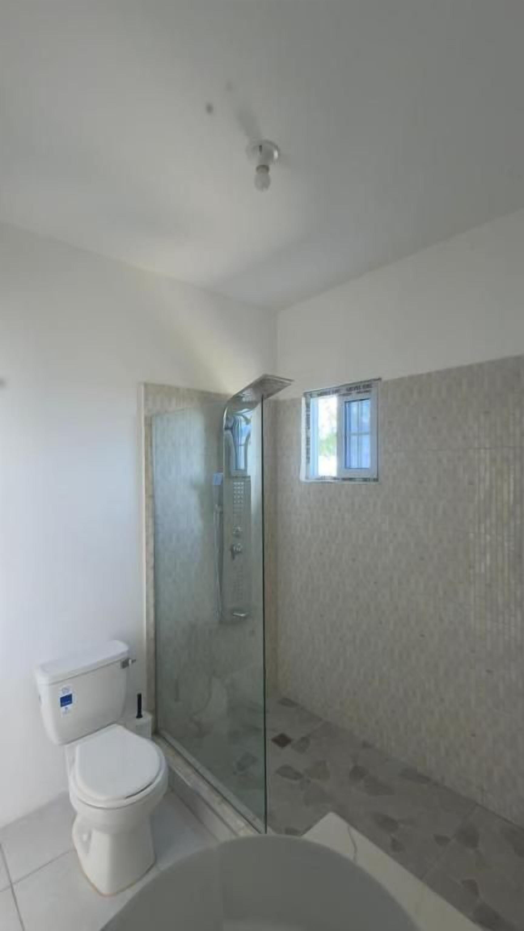 Un baño con inodoro, ducha con mampara de vidrio y una ventana pequeña.