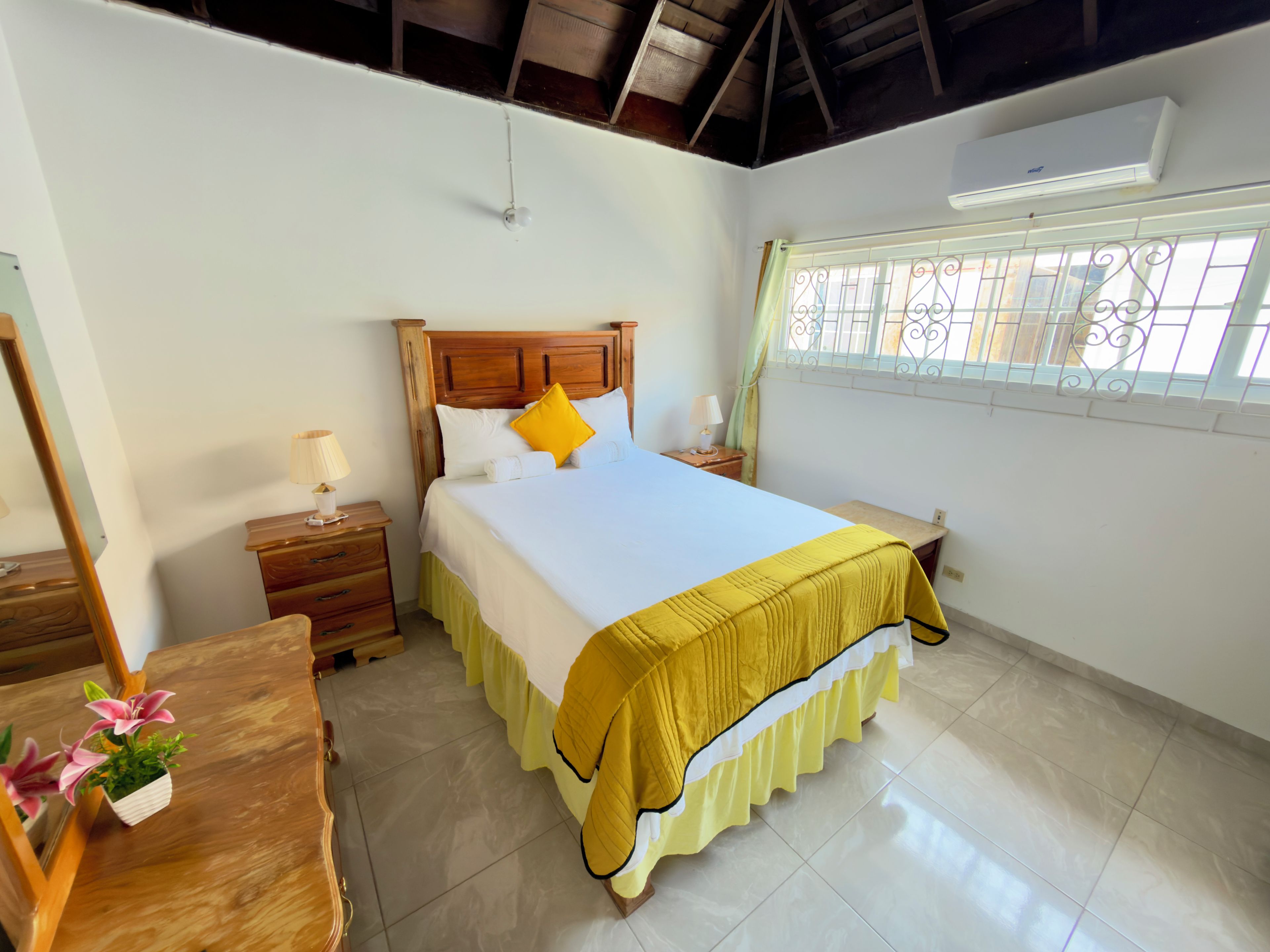 Un dormitorio con cama, mesitas de noche, espejo, ventana con celosías y ventilador de techo.