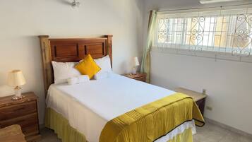A bedroom with a wooden headboard, a bed with white bedding and a yellow pillow, a bedside table with a lamp, and a window with decorative bars.