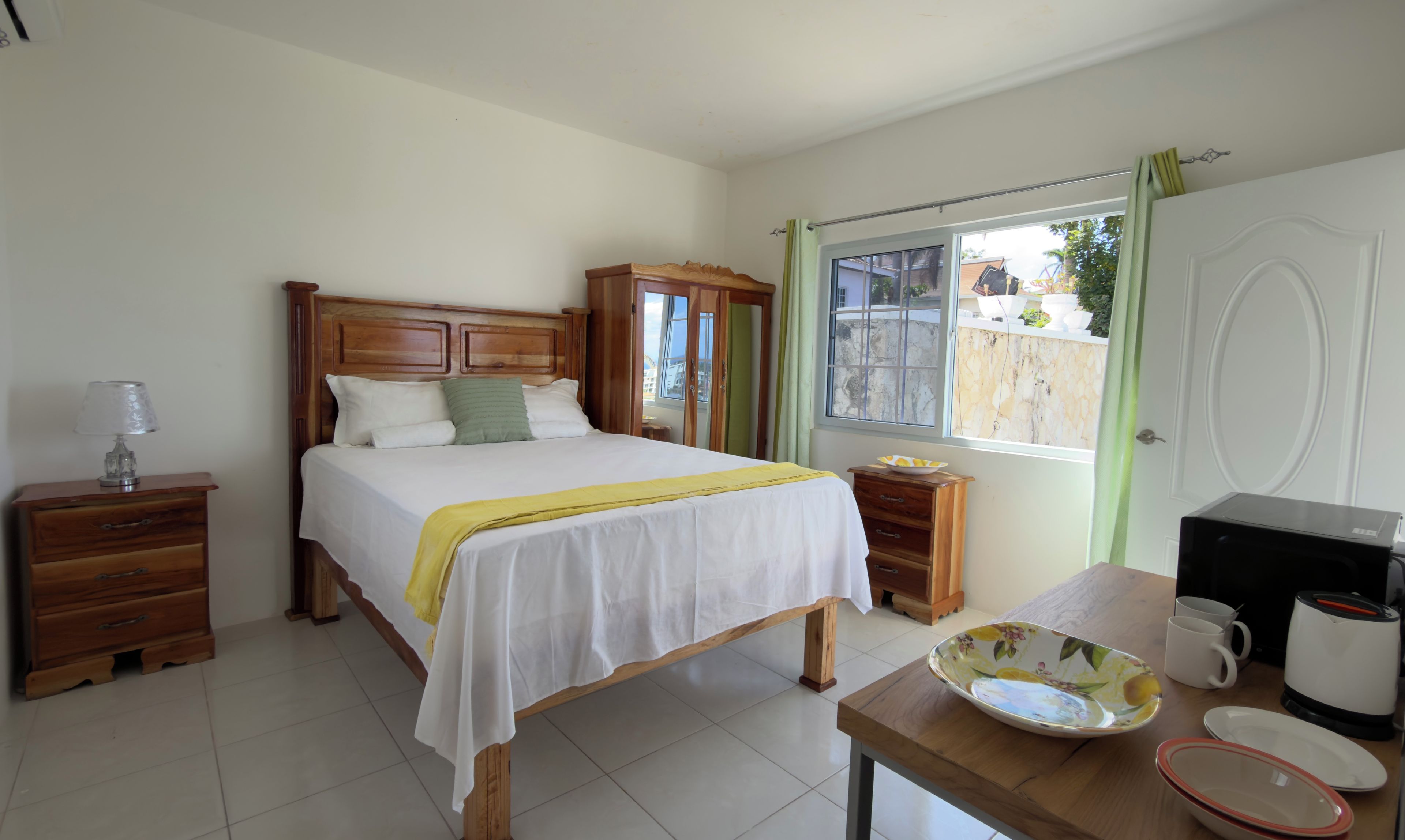 Un dormitorio con cama de madera, una mesita de noche con lámpara, un pequeño aparador de madera, una ventana con vistas y una mesa con tetera y tazas.