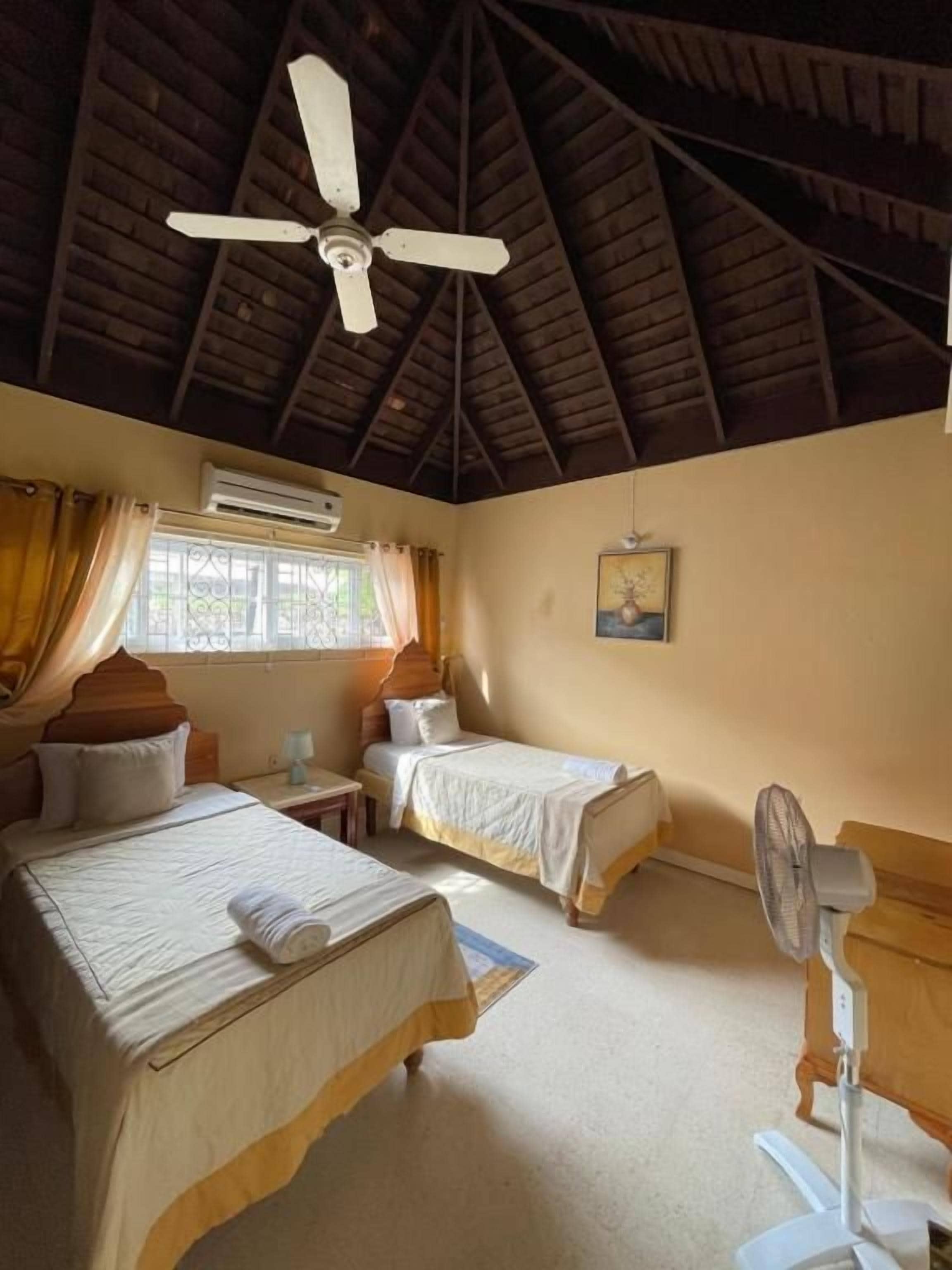Una habitación con dos camas, ventilador de techo, unidad de aire acondicionado y una ventana con cortinas.