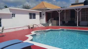 A swimming pool with lounge chairs, a covered patio, and a white building with a red roof.