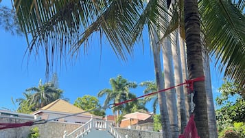 Uma rede sob uma palmeira, com uma escadaria de pedra e uma casa ao fundo.