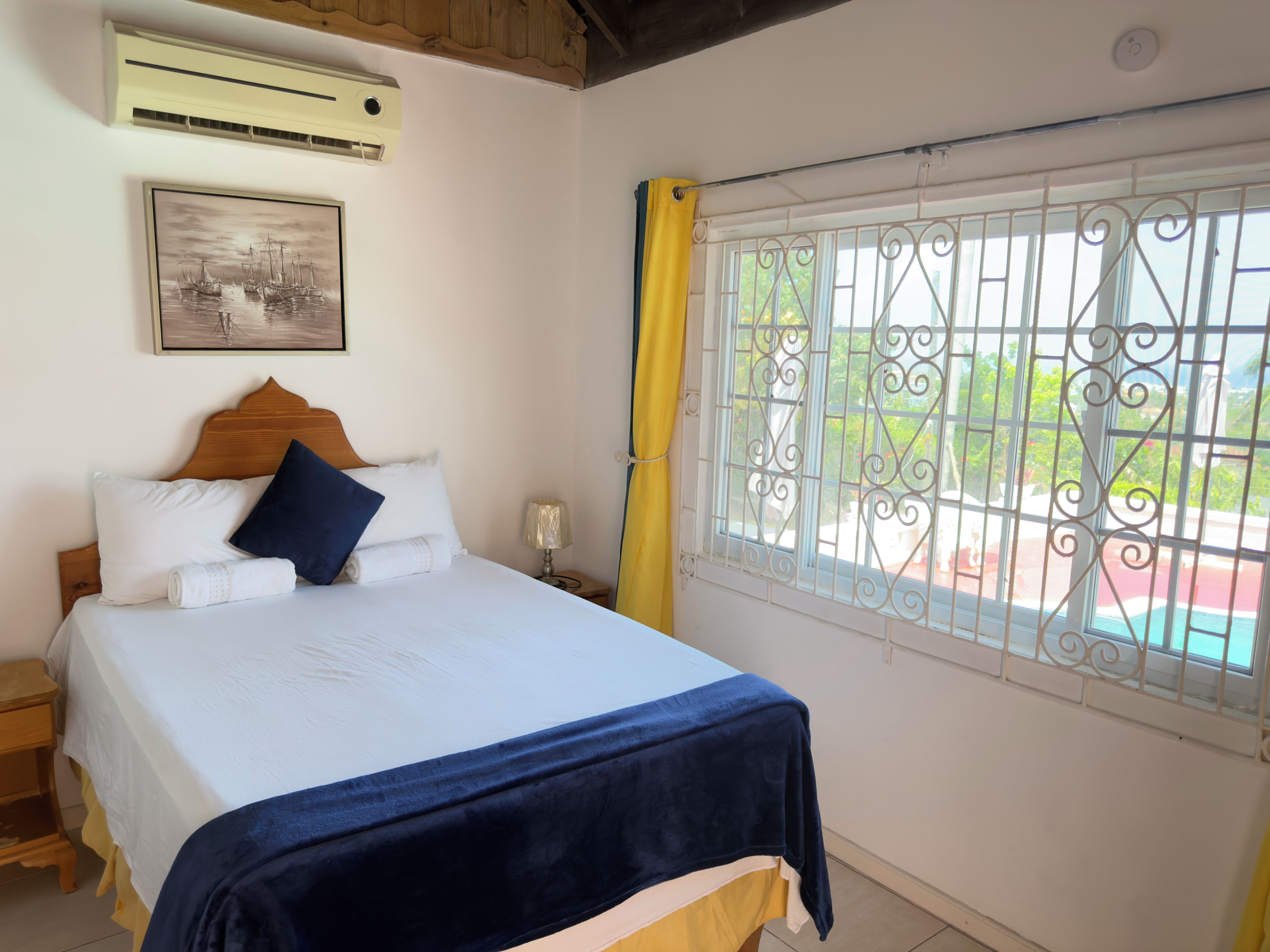 Ein Schlafzimmer mit Bett, Nachttisch, Lampe, einem Fenster mit Verzierungen und einer Klimaanlage.