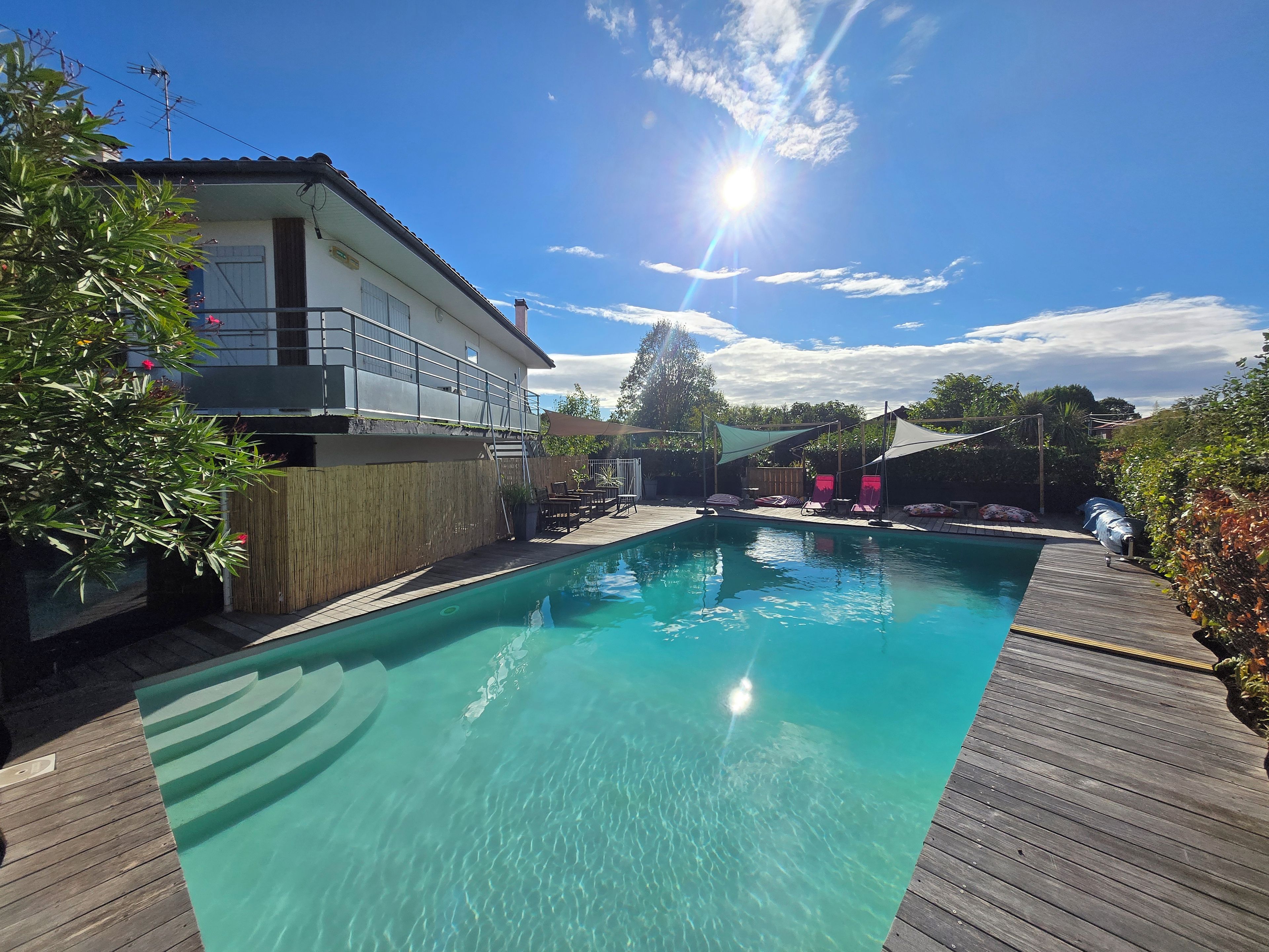 Une piscine aux eaux cristallines, dotée d’une terrasse en bois, entourée d’un espace clos comprenant des chaises longues et un coin ombragé.