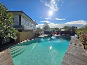 Une piscine aux eaux cristallines, dotée d’une terrasse en bois, entourée d’un espace clos comprenant des chaises longues et un coin ombragé.
