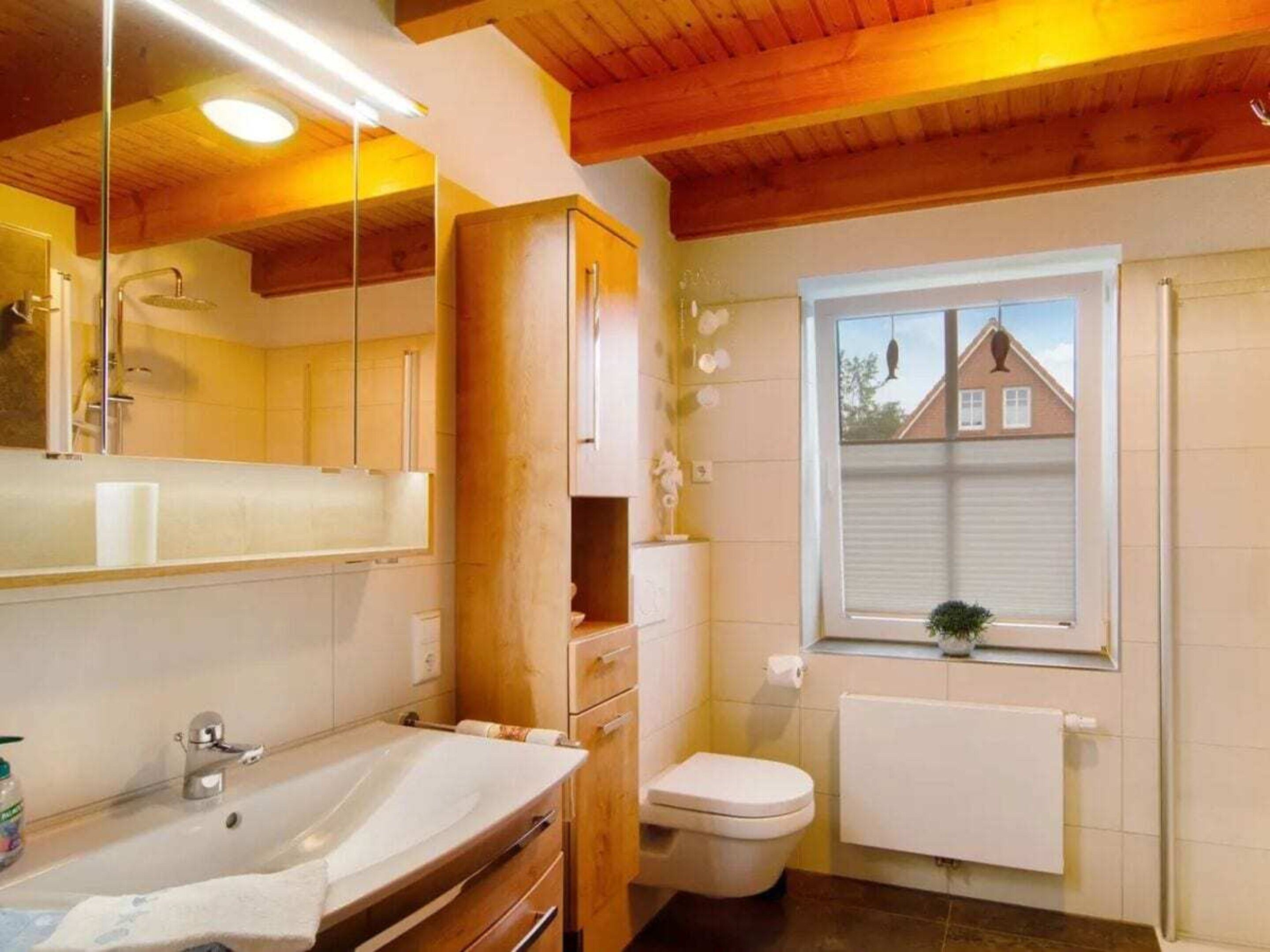 A bathroom with a wooden ceiling, a large mirror, a sink, a toilet, and a window with a view of a house.