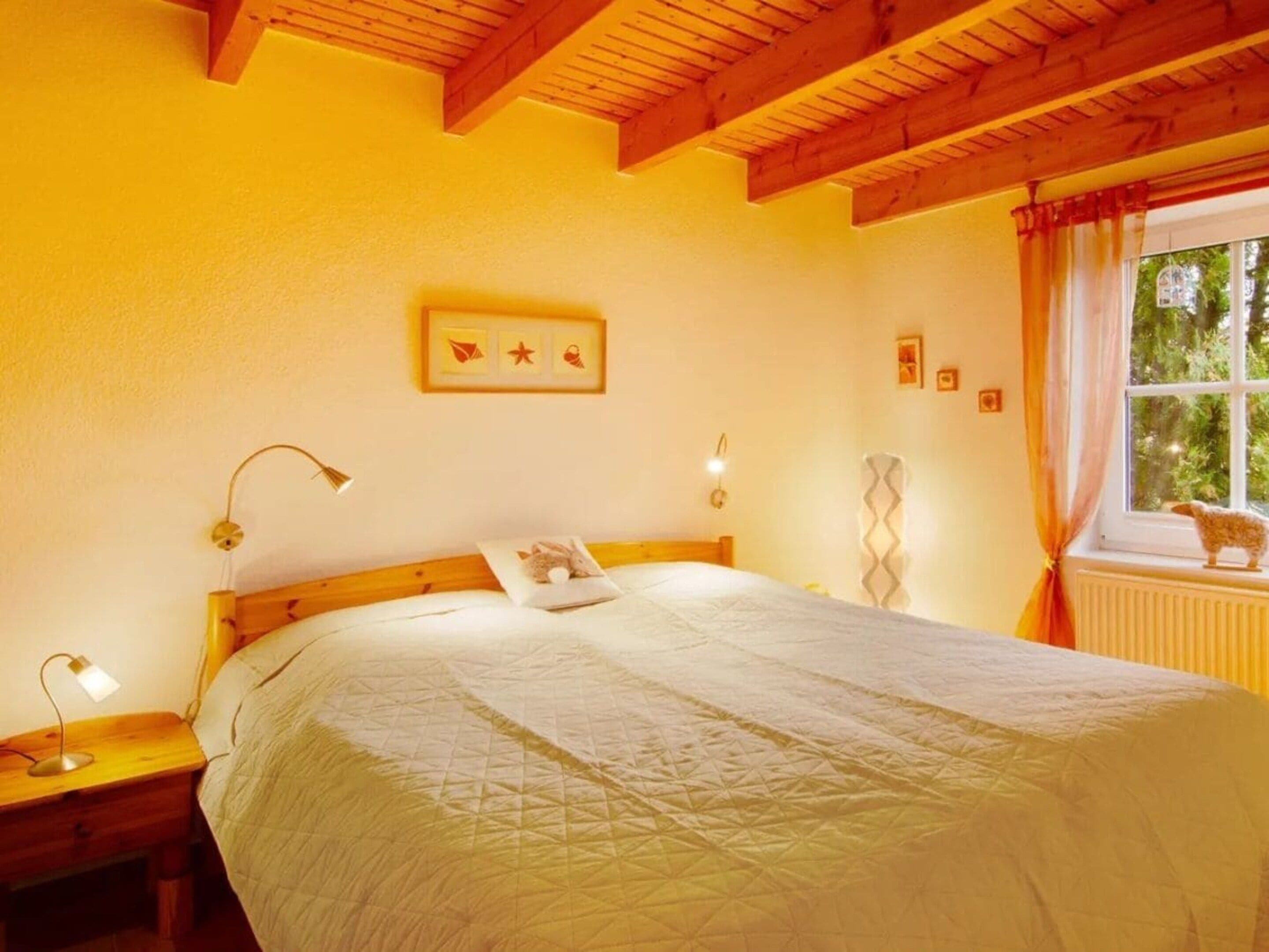 A bedroom with a wooden ceiling, a bed with a white quilt, a nightstand with a lamp, a window with curtains, and a small animal figurine on the radiator.