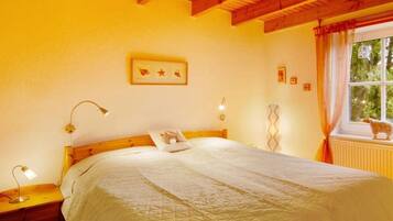 A bedroom with a wooden ceiling, a bed with a white quilt, a nightstand with a lamp, a window with curtains, and a small animal figurine on the radiator.