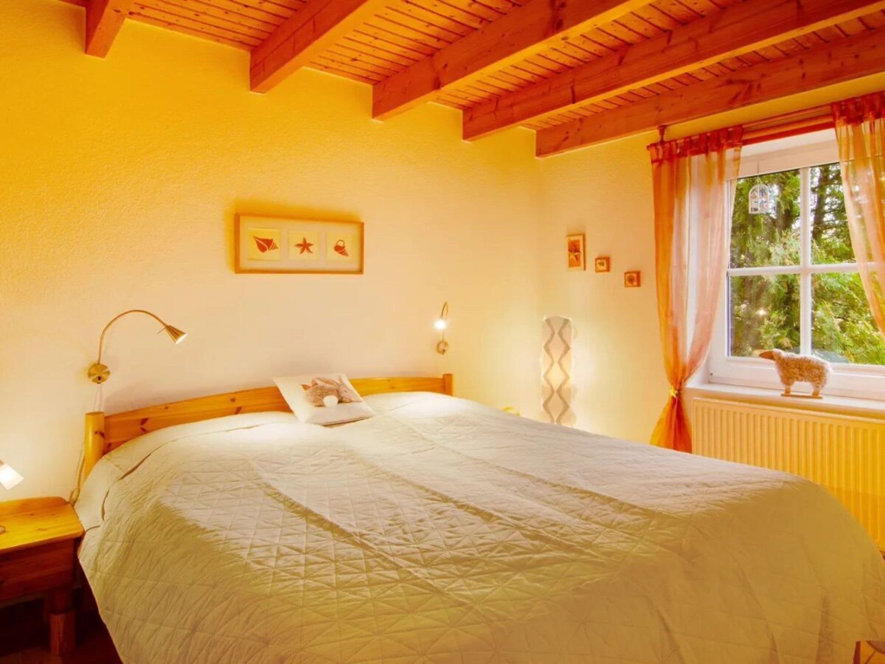 A bedroom with a wooden ceiling, a bed with a quilt, a bedside lamp, a window with curtains, and a small animal figurine on the windowsill.