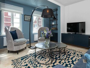 Ein Wohnzimmer mit rundem Couchtisch, zwei Sesseln, einer Stehlampe, einem Fernseher und einem Bücherregal.