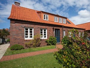 Ein zweigeschossiges Backsteinhaus mit rotem Ziegeldach, einer Haustür und einem kleinen Vorgarten mit Rasen und Sträuchern.