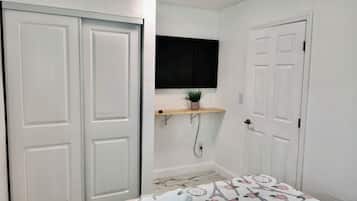 Quarto com cama, televisão na parede, prateleira com planta e uma porta.