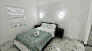 A bedroom with a bed, a nightstand, a window with blinds, and a door.
