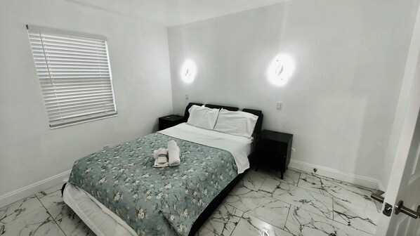 Un dormitorio con cama, mesita de noche, ventana con persianas y puerta.