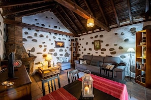 Ein rustikales Interieur mit einer Steinsandsteinmauer, Holzbalken, Kamin, einer Couch, einem Esstisch und einem Bücherregal.