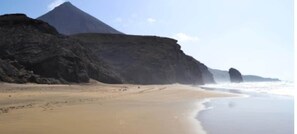 A beach with a large rock formation and waves crashing onto the shore. - Welcome (Puerto del Rosario)
