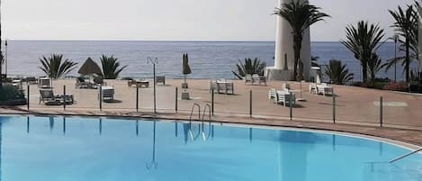 Un área de piscina con sillones de descanso y sombrillas, una alta columna blanca y vista al mar.