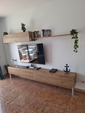 A modern living room with a flat-screen TV mounted on a wooden console, a bookshelf above it, and a hanging plant to the right. - Vv Casa Josué (1 GB Internet) (Castillo Caleta de Fuste)