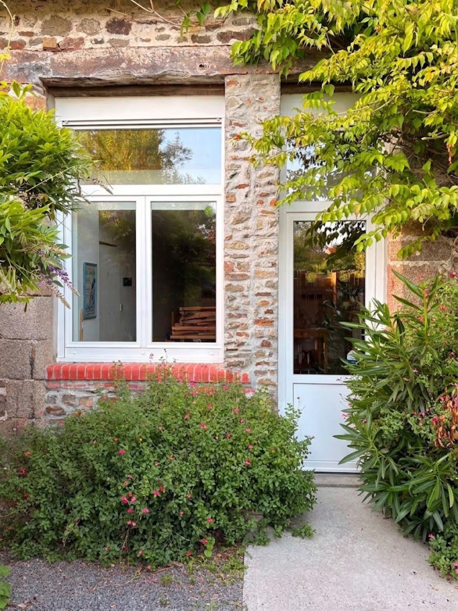 A stone building with a white door and windows, surrounded by greenery and a gravel path.