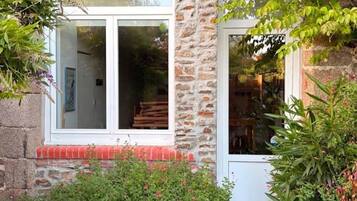 A stone building with a white door and windows, surrounded by greenery and a gravel path.