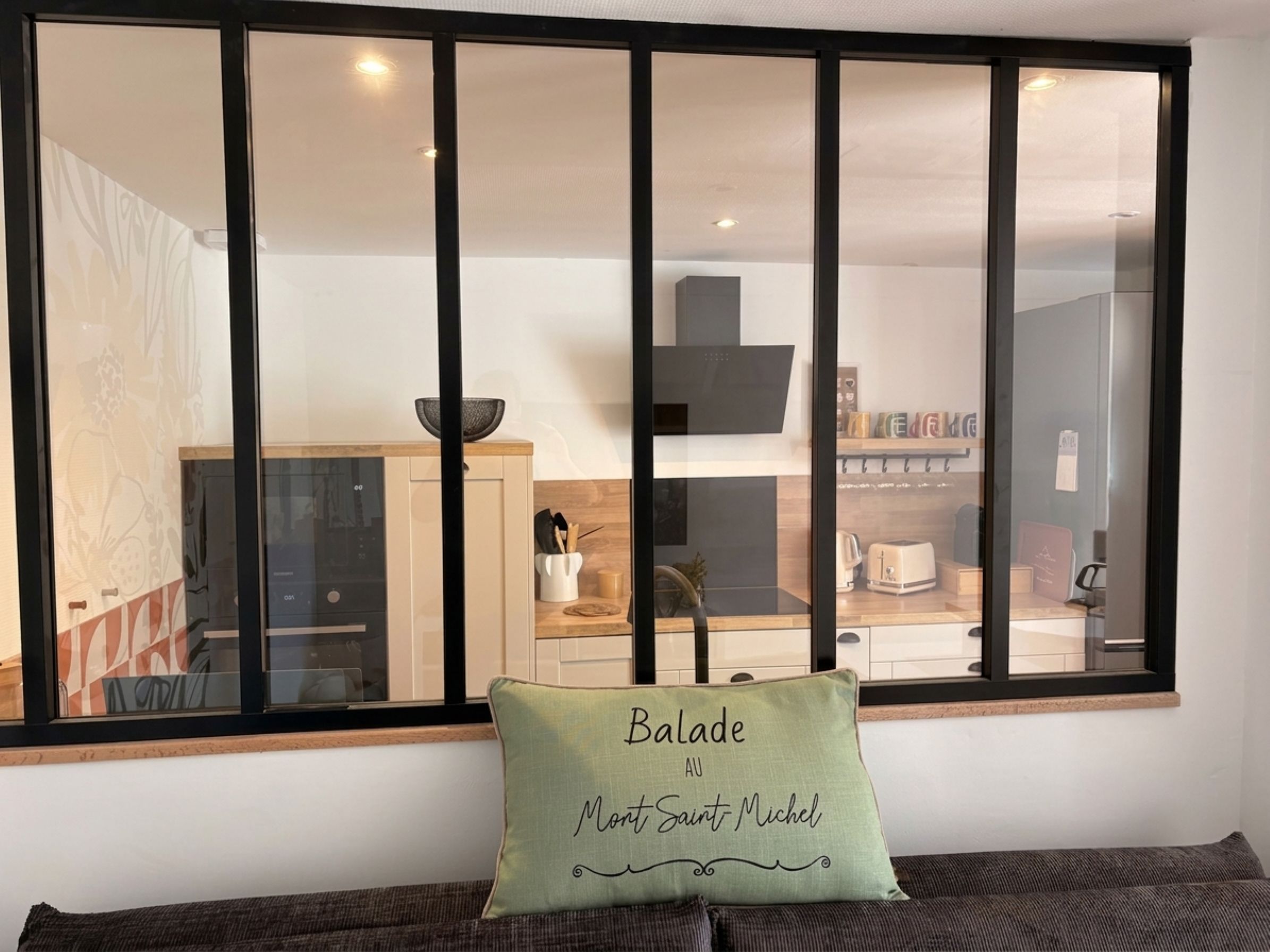 A room with a large window, a green pillow with text, and a view of a kitchen area.
