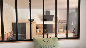 A room with a large window, a green pillow with text, and a view of a kitchen area.